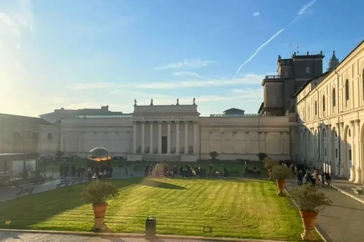 Pátio interno dos Museus do Vaticano com visitantes caminhando entre jardins e edifícios históricos.